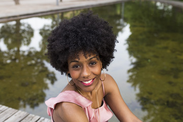 portrait of  a Happy young beautiful afro american woman sitting on wood floor and smiling. water background. Spring or summer season. Millennial. Casual clothing.