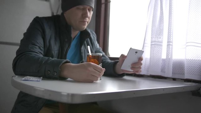 A young man drinks tea from a special glass cup in a train, looks into his white smartphone, looks at the train window, pushing the curtains, Russia