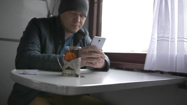 A young man in a gray hat and blue jacket goes on a train reading information in his white smartphone, drinking tea from special glass cups in a train