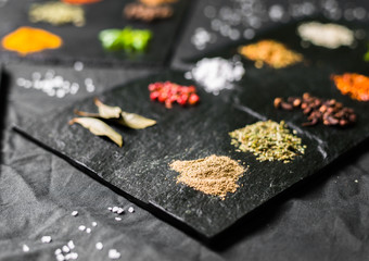 Large selection of different colorful contrast spices and seeds on slate against black background