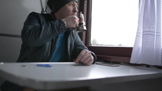 A young man in a gray hat and a blue vest goes on a train, drinking tea from special glass cups in a train, looks out the train window, reading information in his white smartphone