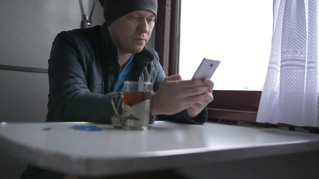 A young man in a gray hat and blue jacket goes on a train holds in his hands and communicates on his white smartphone, smiles, puts the smartphone on the table and takes a glass of tea