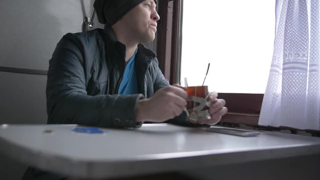 A young man in a gray hat and a blue vest goes on a train, drinking tea from special glass cups in a train, looks out the train window