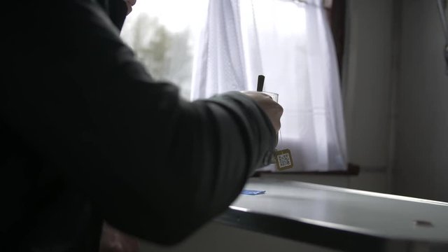 A young man rides a train in a dark jacket, sits at a table, drinks tea from a special glass cup, against the background of a window with a white curtain. Close-up, side view