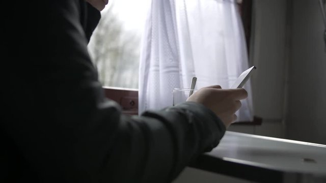 young man goes on a train, sits at a table, takes a smartphone in his hands, turns off his smartphone takes a glass of tea, against the window with a white curtain. Close-up, side view