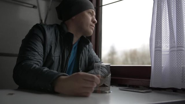man is traveling on a train, sitting at a table, pulling back the curtain, looking out the window, drinking tea from a special glass, taking his smartphone, is in correspondence, smiling