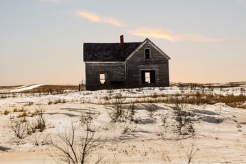 horizontal image of an old abandoned house with dead grass and snow all around with the sun starting to set in the early evening.