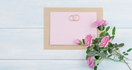 Pink roses and bridal rings with paper greeting card for wedding on background of shabby wooden planks.