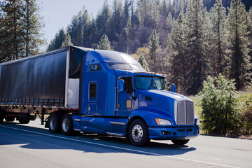 Big modern famouse rig semi truck blue and black trailer tented on road in forest hills