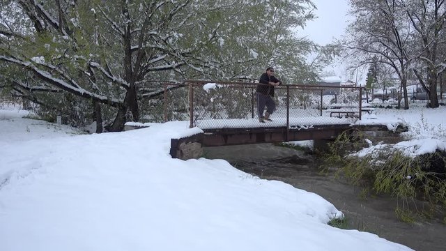 A man looks out at a running stream in the winter and playfully throws snowballs.  	