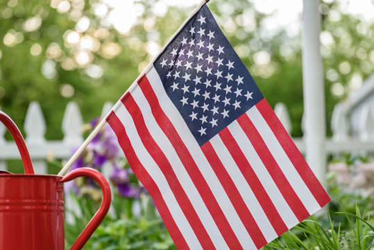 American flag in red watering can with white picket fence in background
