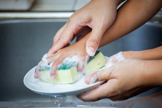 Mother And Child Hand Washing Dishes Over The Sink In The Kitchen Together