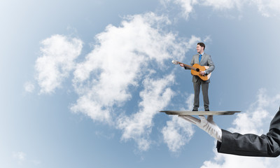 Businessman on metal tray playing acoustic guitar against blue sky background