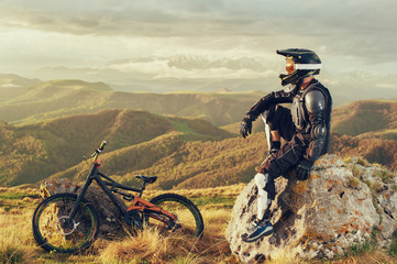 A professional downhill rider sits on a stone resting after a tense race on a mtb bike