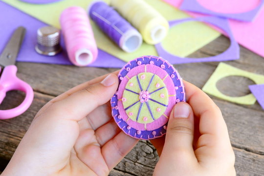 Small child made a beautiful flower from felt circles and beads. Child holds a felt flower in hands. Felt flower DIY, craft supplies on wooden table. Easy and fun sewing. Teaching kids to sew by hand