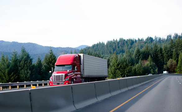 Red contemporary semi truck cargo reefer beside concrete fence devided lines highway evergreen trees