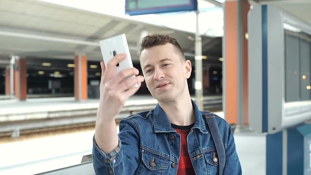 Handsome man in denim jacket doing selfies on smartphone on the train station, steadycam shot
