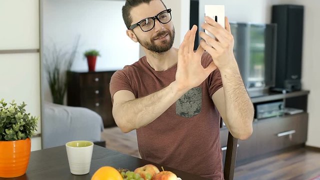 Handsome man sitting in the flat and doing selfies on smartphone, steadycam shot
