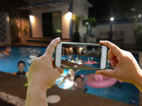 Take a group picture of blurred people on swimming pool background with hand holding smartphone in the fun partty on night