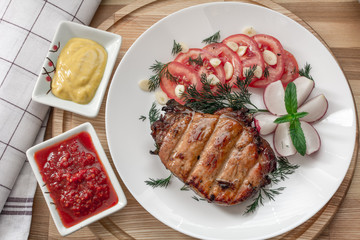 Pork steak cooked on the grill is decorated with a tomato, radish, dill, parsley, greens garlic mint, adjika and mustard.