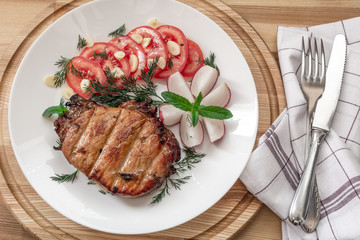 Pork steak cooked on the grill is decorated with a tomato, radish, dill, parsley, garlic greens, mint with a fork and knife on a tissue napkin on a wooden background