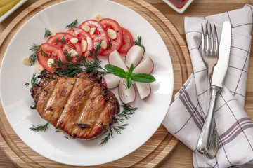 Pork steak cooked on the grill is decorated with a tomato, radish, dill, parsley, garlic greens, mint with a fork and knife on a tissue napkin on a wooden background