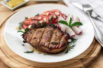 Pork steak poured with sauce is decorated with tomato, radish, dill, parsley, garlic greens, mustard, mint, cutlery on napkin, shallow depth of field