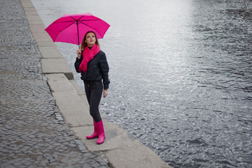 Beautiful young and happy blond woman in a bright pink scarf, rubber boots and umbrella walking in a rainy city.