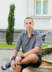 Obraz premium Man sitting on the stone fountain. Young man smile on a summer day in shirt