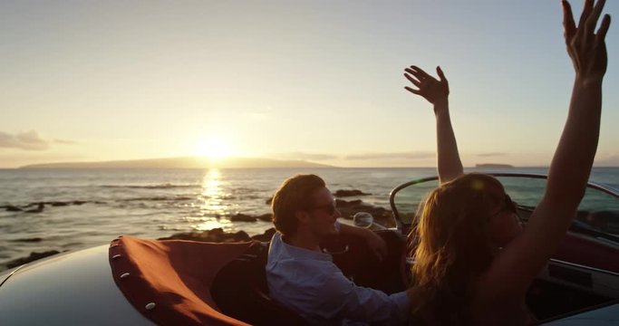 Romantic Couple Hands in thr Air Driving in Vintage Convertible Car at Sunset on Country Road by the Sea
