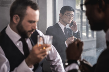 Serious asian businessman smoking cigar and talking on smartphone while colleagues drinking whisky, multicultural business team