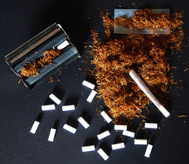 Rolling machine, hand rolled cigarette with tobacco and scattered filters on black table 