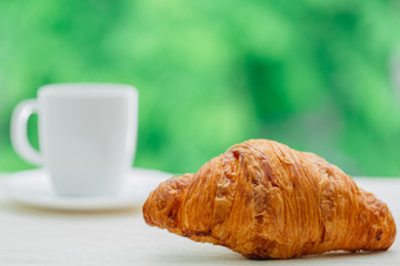 Tasty buttery croissant and cup of coffee on white wooden table over green tree background. Traditional french breakfast