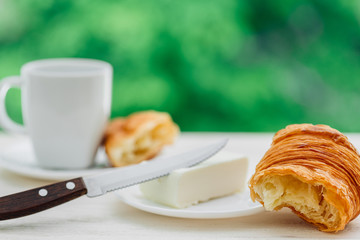 Tasty crunchy croissant with butter and knife and cup of coffee on white  wooden table over green tree background. Traditional french breakfast