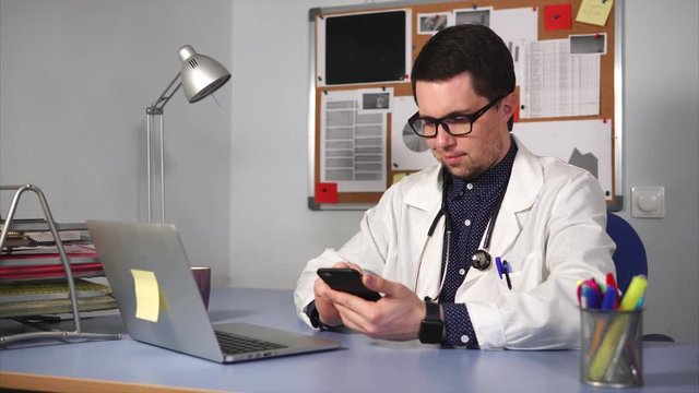 Medical student studying. Male medic in white coat with stethoscope sitting at the desk and using smart watch.