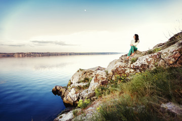 Naklejka premium Young woman sits on rock and enjoys view of river at sunset