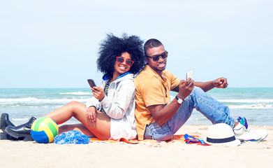 Happy afro american couple using mobile phone back to back on the beach  