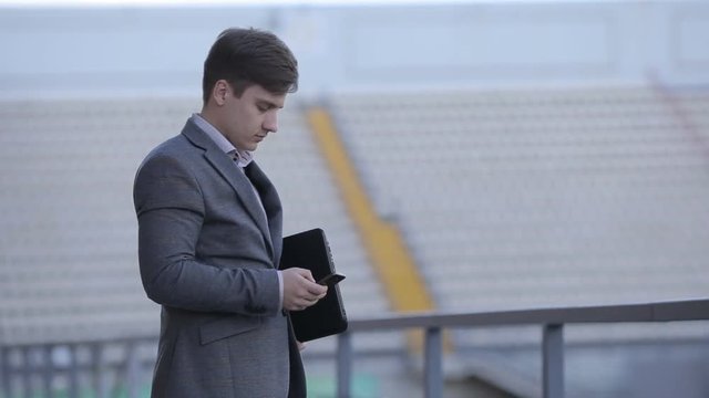 Young man in a business suit standing on the stadium with a gadget in his hand. Man with laptop and phone.