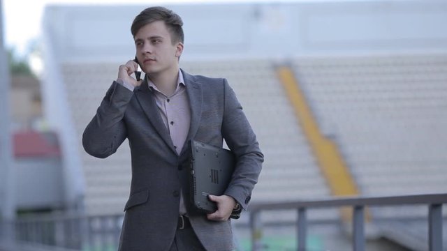 Young man in a business suit standing on the stadium with a gadget in his hand. Man with laptop and phone.