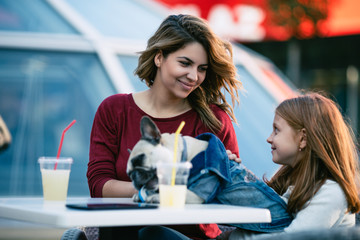 Mother and daughter sitting in cafe restaurant and enjoying with their French bulldog puppy. People and dogs theme.