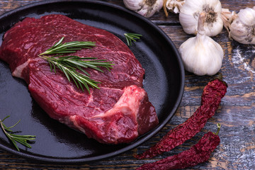 Raw Beef Steak, salt, pepper, garlic, rosemary  on the black pan, wooden background.