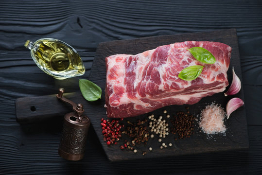 Raw fresh pork neck meat with seasonings on a black wooden chopping board, elevated view, studio shot