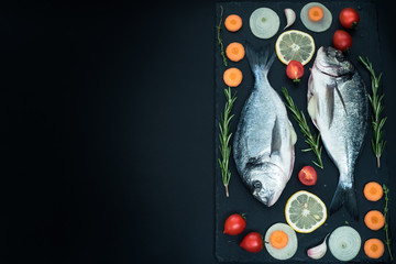 Fresh raw uncooked dorado fish with lemon, herbs, oil, vegetables and spices on black backdrop, top...