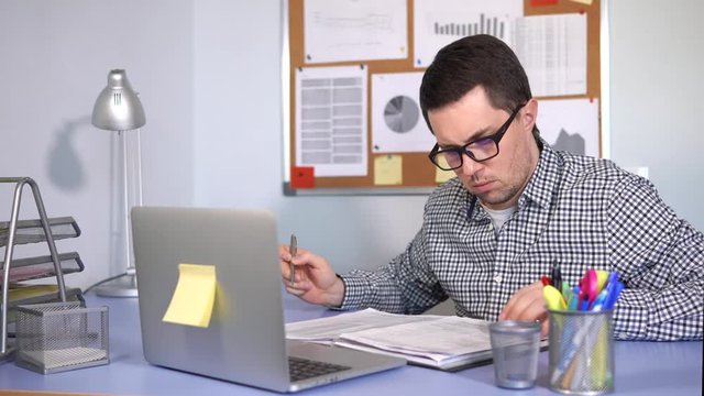 Manager fills in the reporting documentation for his firm. A person with poor eyesight makes a marking pen on paper, on his desktop is a laptop.