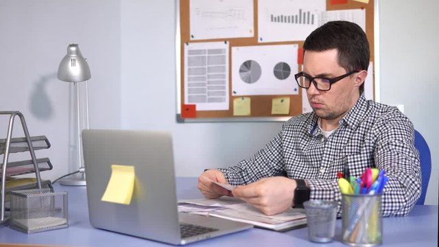 The head of a small firm checks the documents with a serious look, emphasizing the important points. A man is sitting in the office at the desk with a laptop.