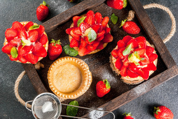 Summer and spring dessert. Home pies tartlets with custard and strawberries, decorated with mint and powdered sugar. On black stone table, rustic, with wooden board, tray. Copy space