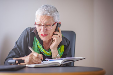 Happy senior old business woman writing down orders, talking on her cell phone