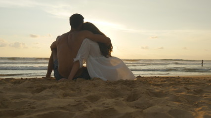 A woman and a man sits together in the sand on the sea shore, admiring the ocean and landscapes. Young romantic couple is enjoying beautiful sunset sitting on the beach and hugging. Close up