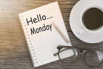 Concept hello monday message on notebook with glasses, pencil and coffee cup on wooden table.