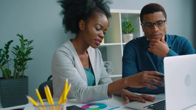 Two african american business people working together at the office. Afro American businessman and businesswoman talking, using laptop computer, checking financial charts. Team work